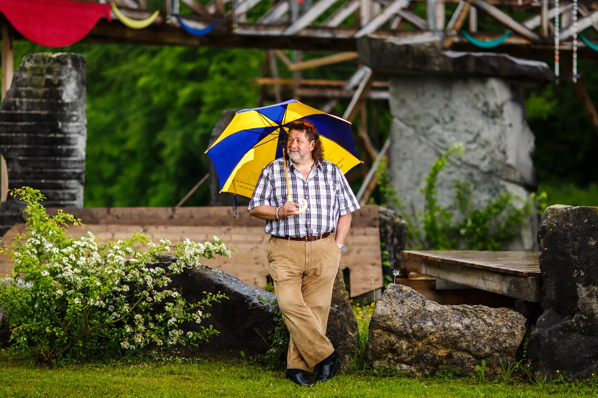 Ein bärtiger Mann mit langen Locken steht unter einem gelb-blauen Regenschirm.